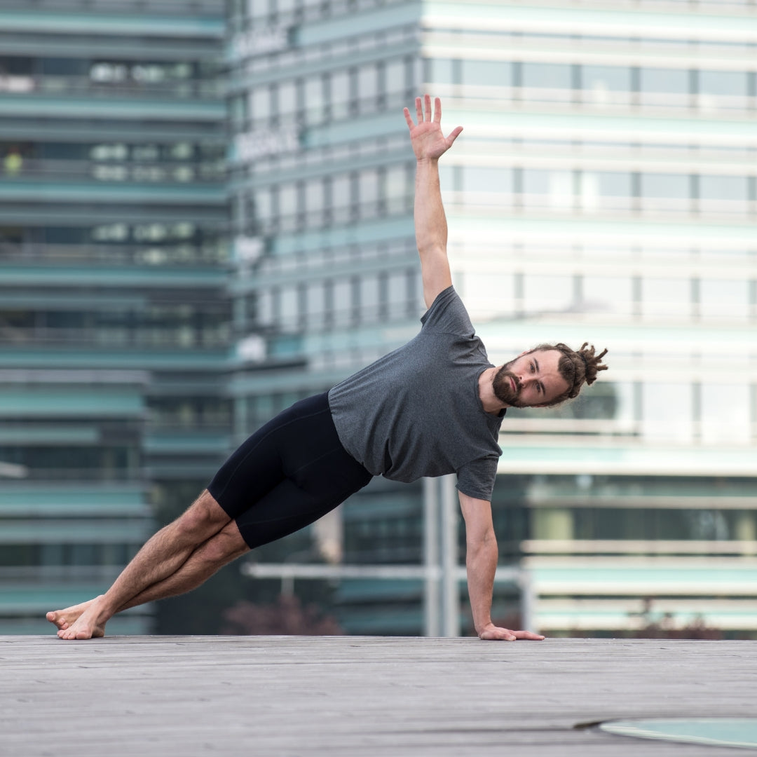 A man in a gray t-shirt and black shorts performs a side plank yoga pose on a concrete rooftop with a blurred modern glass building in the background.